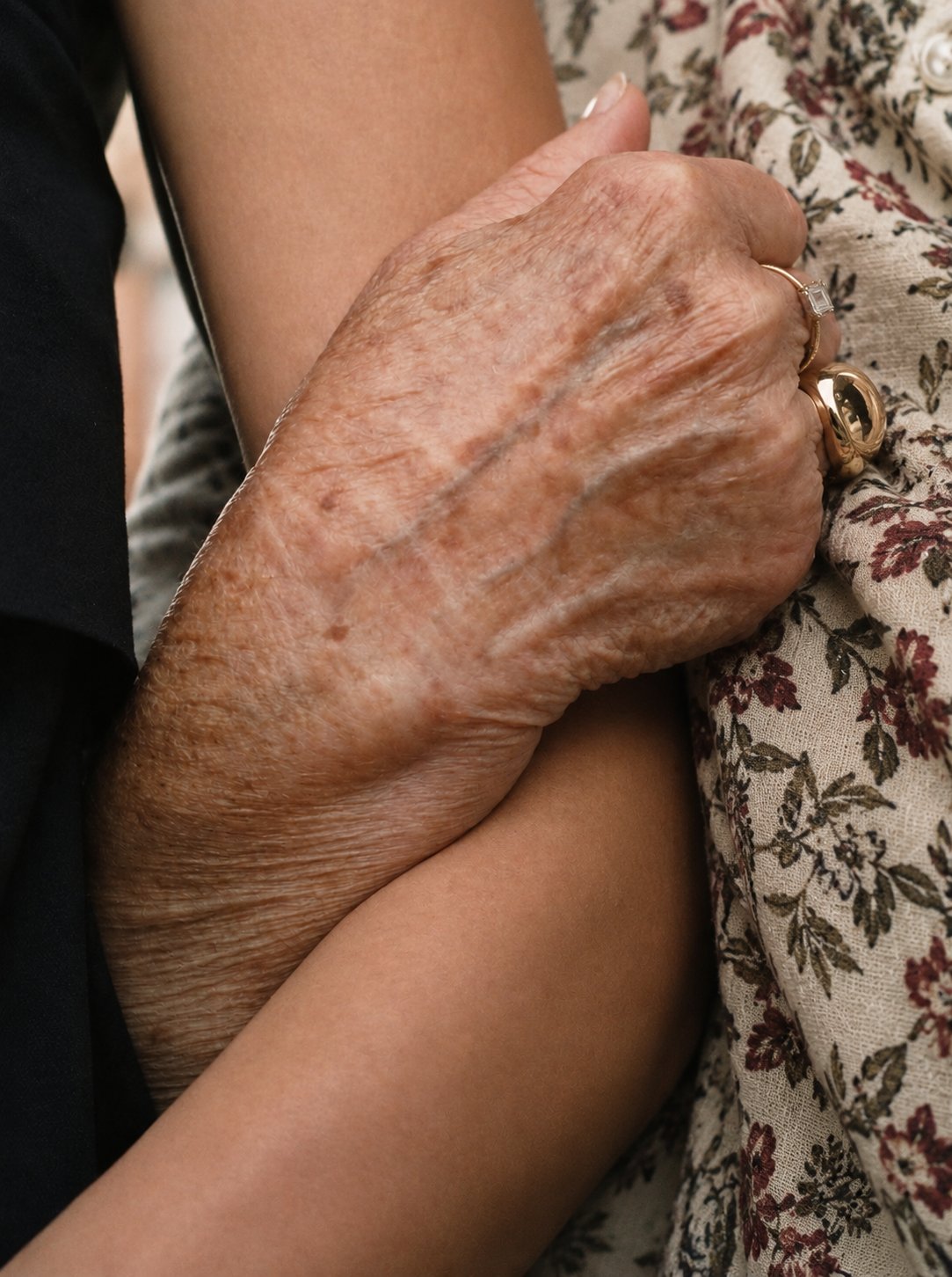 An older hand with gold rings resting on a younger person's forearm